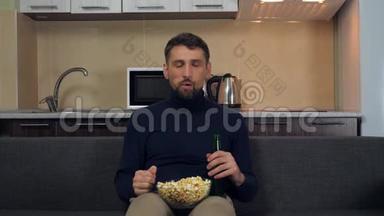 一个穿着深蓝色套衫和灰色裤子的年轻人坐在沙发上，吃爆米花，喝啤酒，看电视上的<strong>娱乐节目</strong>